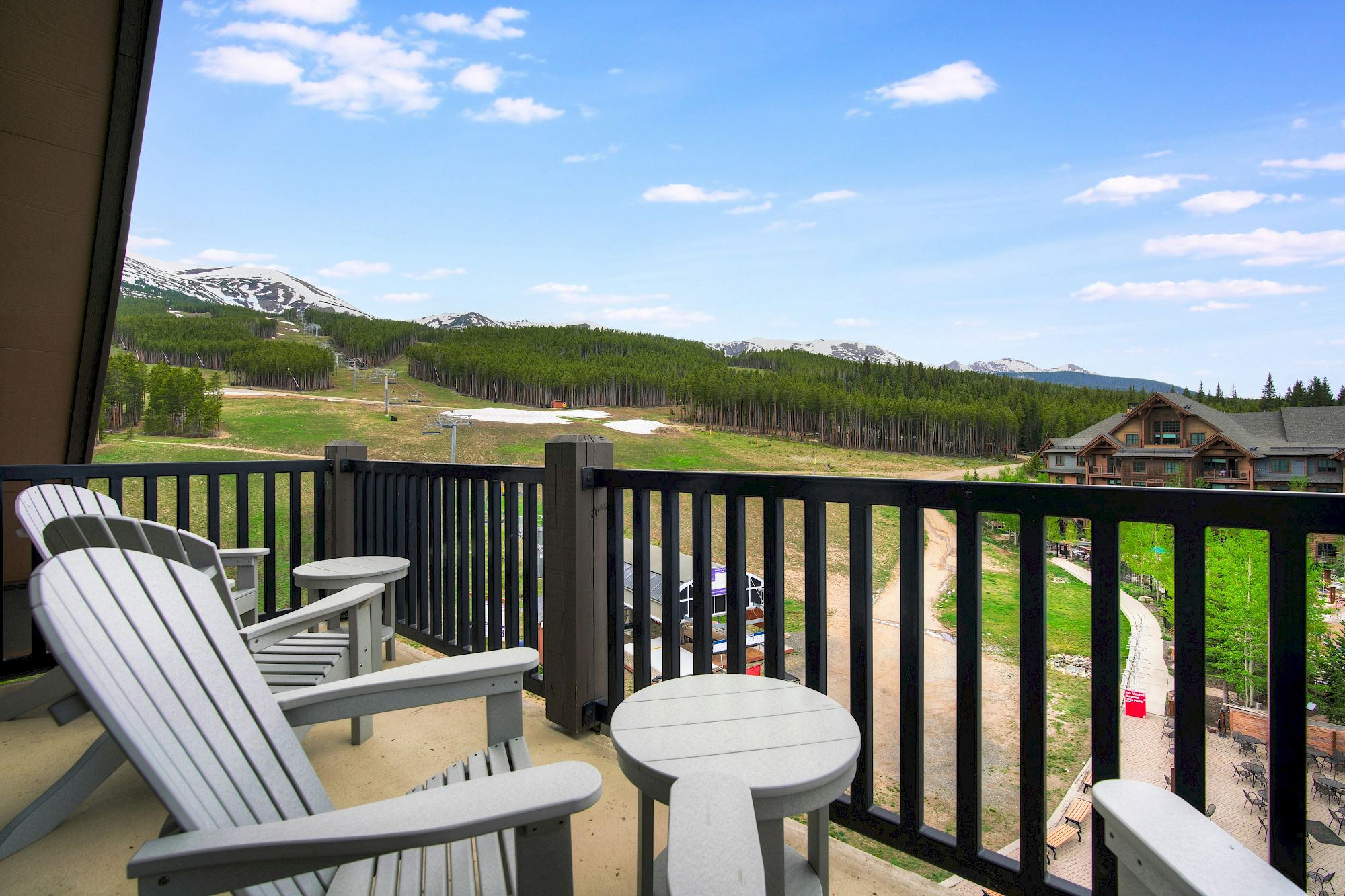 A mountain ski resort view from a balcony, with Adirondack chairs, winding paths, lush green slopes, and snow-dusted peaks under blue skies.