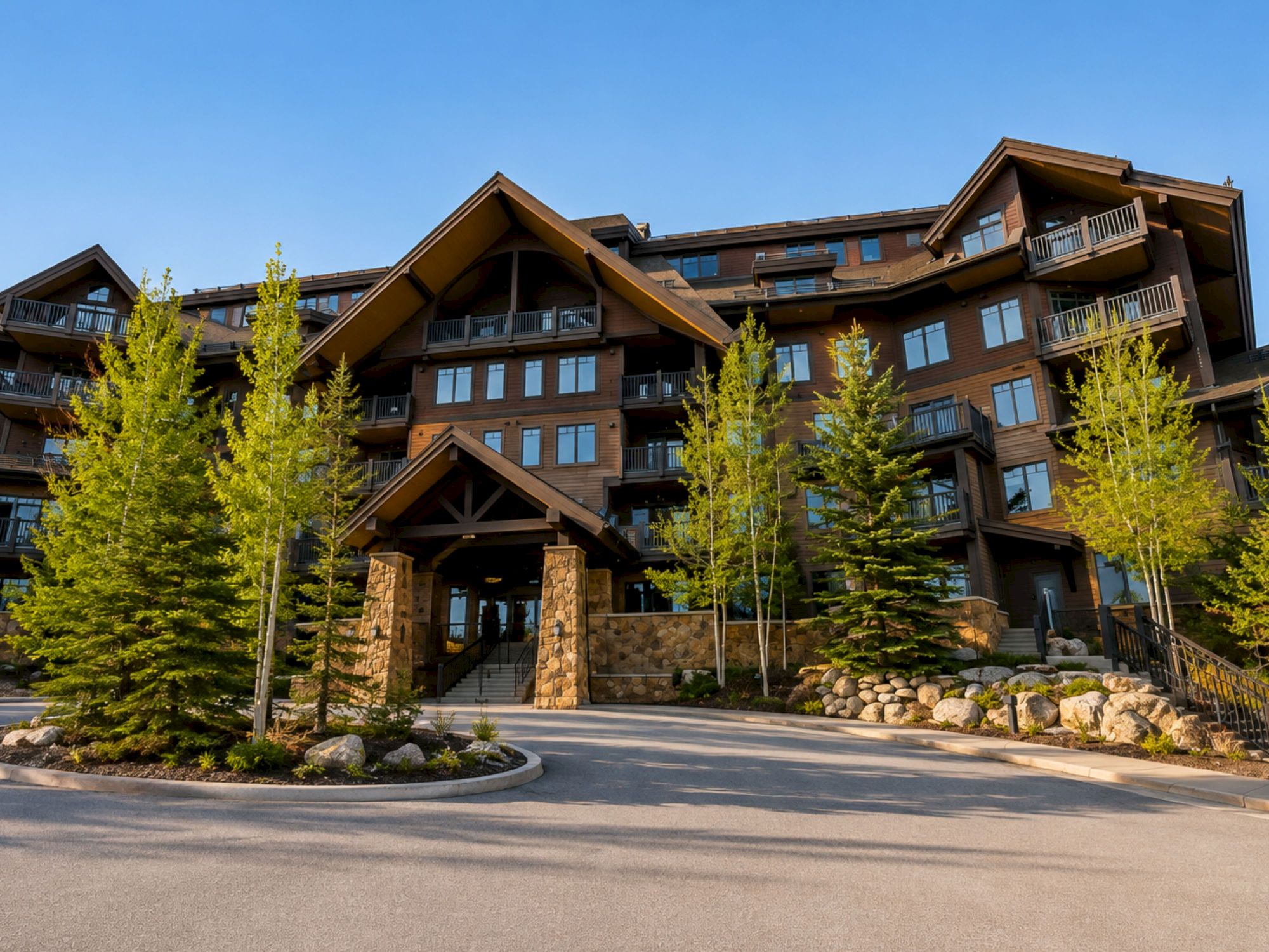 A modern apartment complex with wood-and-stone balconies, framed by trees and landscaped walkway under a clear blue sky.