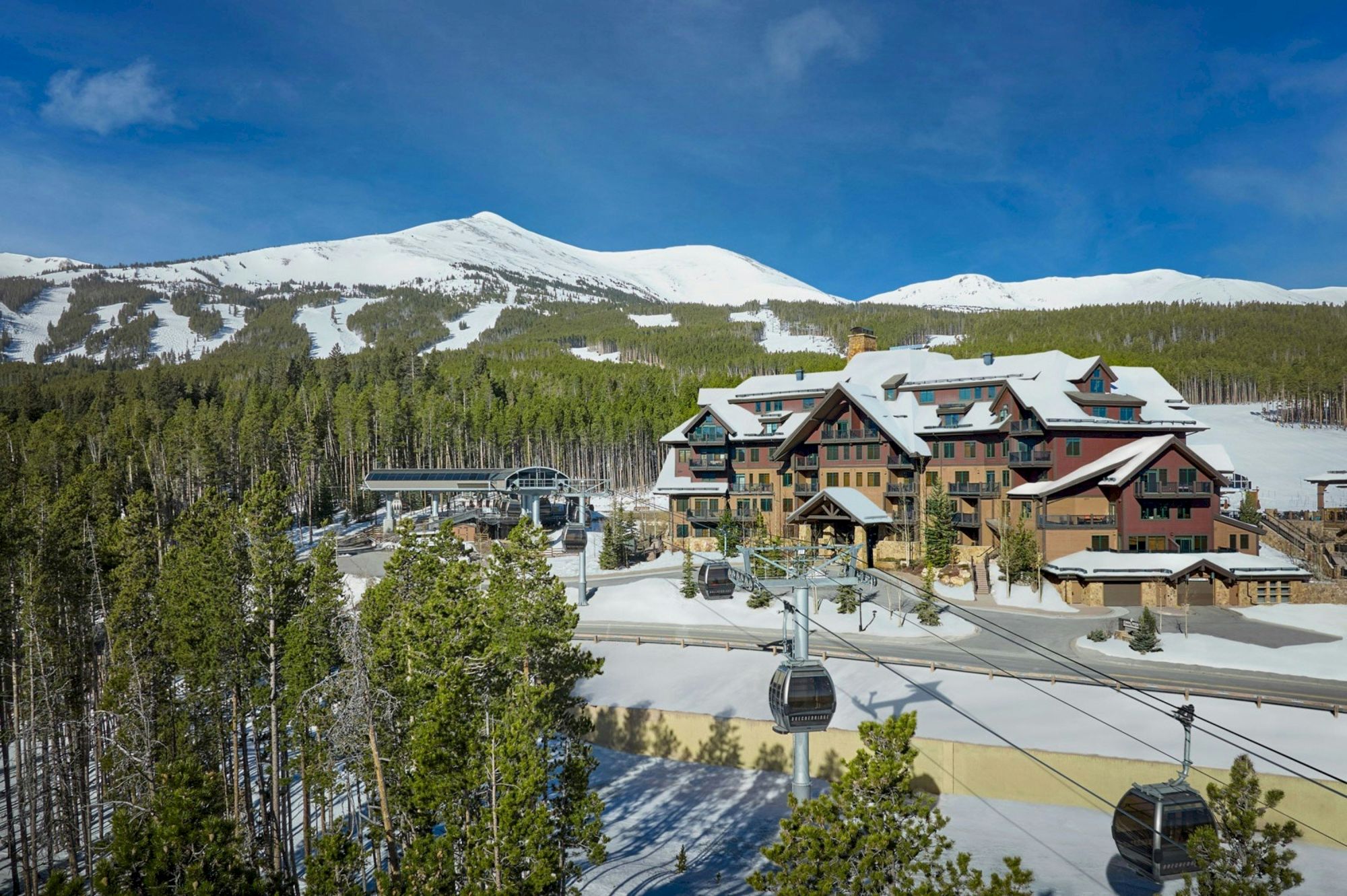 A snowy ski resort lodge sits amid pine trees, with gondola cables and white mountains under a blue sky.