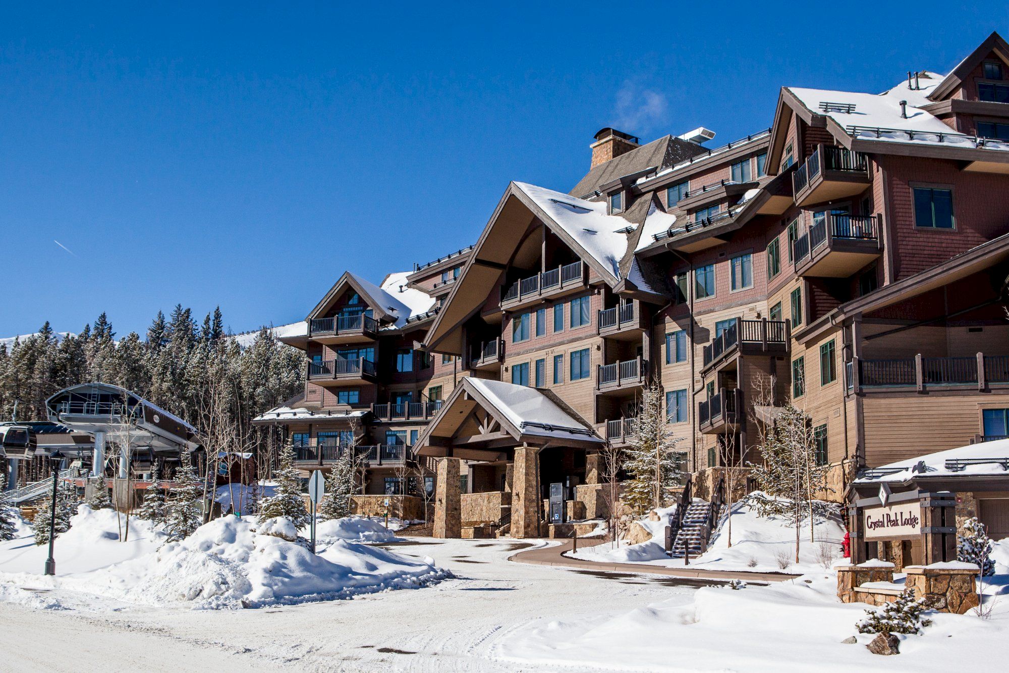A snowy mountain lodge with multi-story balconies and pitched roofs under a clear blue sky, with &ldquo;Crystal Peak Lodge&rdquo; signage visible.