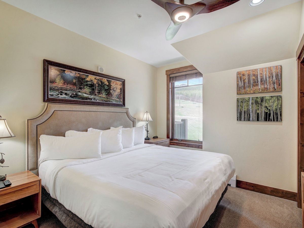 A bright bedroom with a large bed, ceiling fan with lights, two landscape wall artworks, and big windows showing a forest view outside.