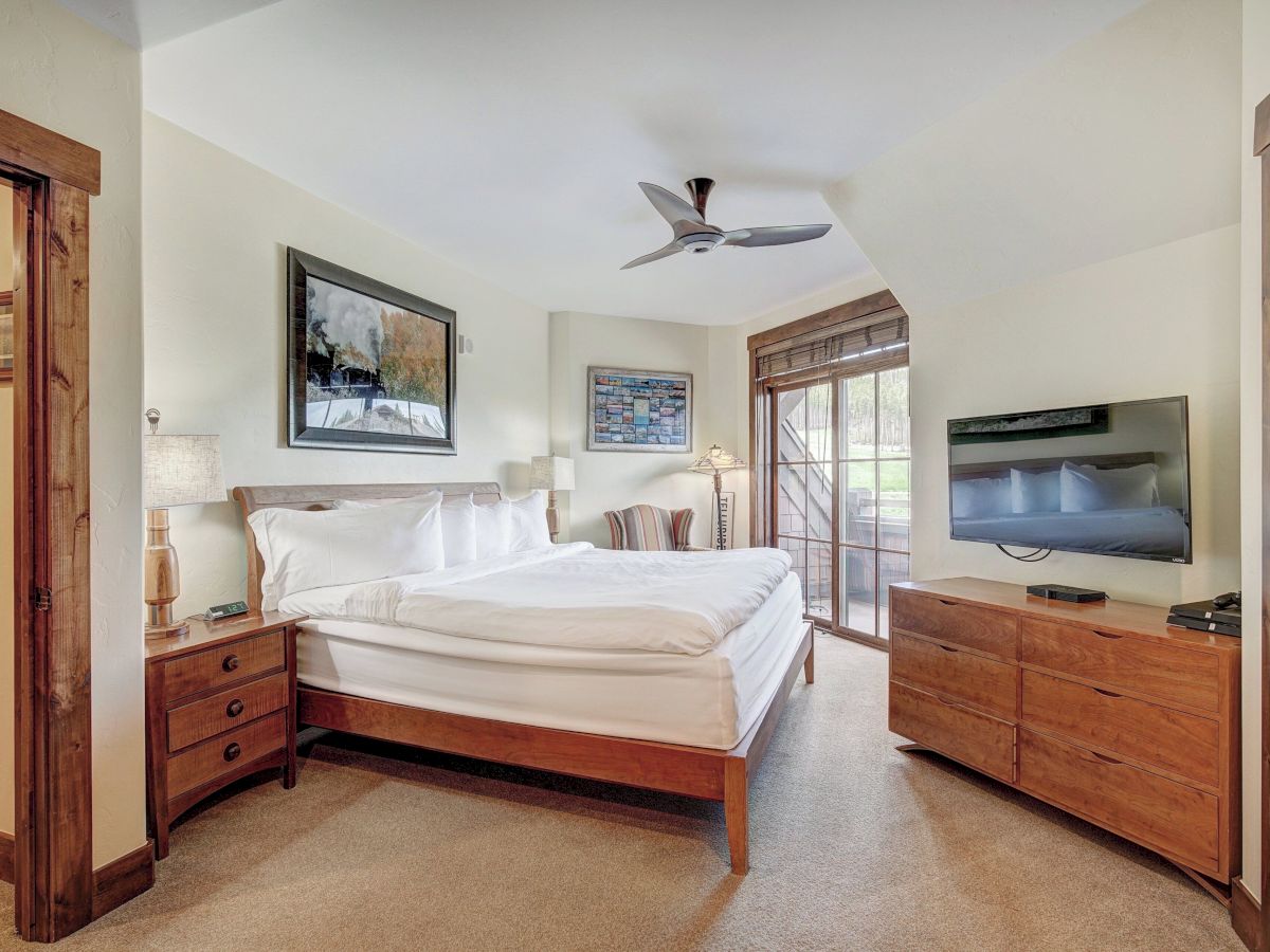 A bright bedroom with a wooden bed, ceiling fan, wall art, big TV on a dresser, and windows with blinds.
