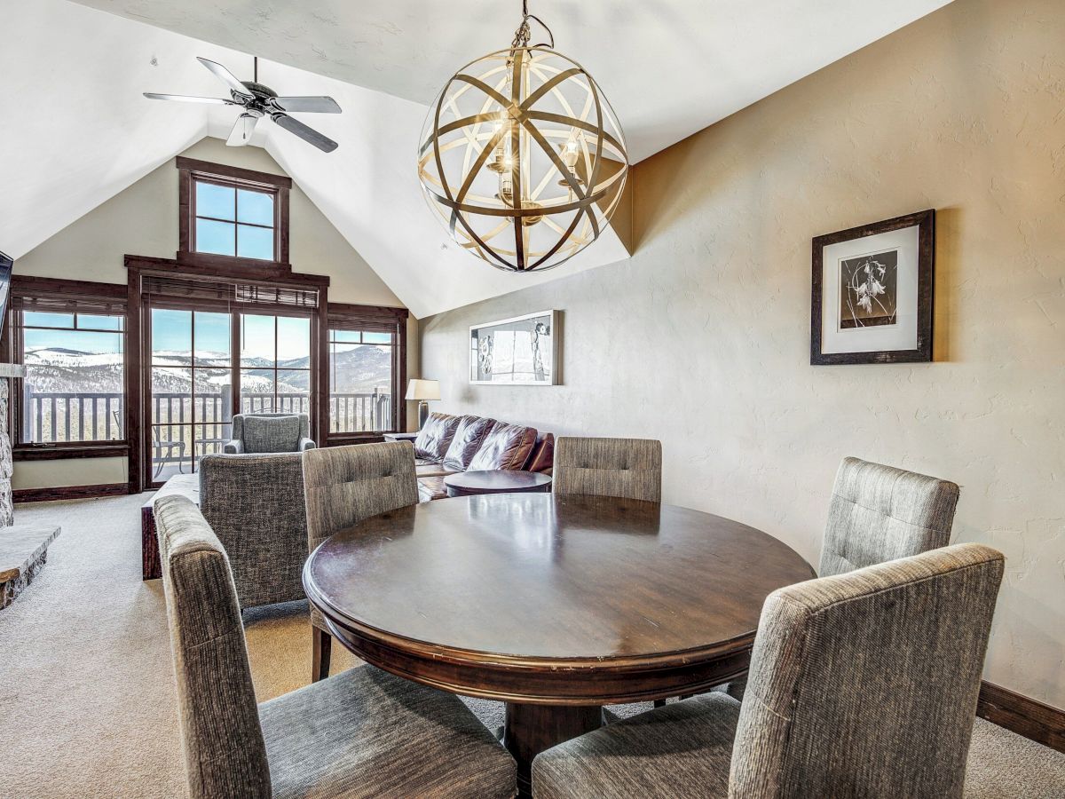 Bright living room with a round wooden table, cushioned chairs, hanging chandelier, fireplace, and large mountain-view windows.