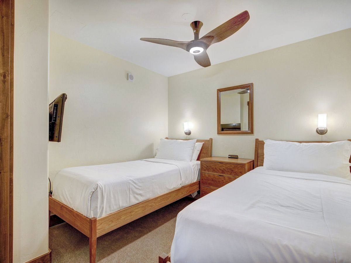 A bright hotel-style bedroom with two twin beds, wooden headboards, a ceiling fan, and bedside lights with a wall mirror.