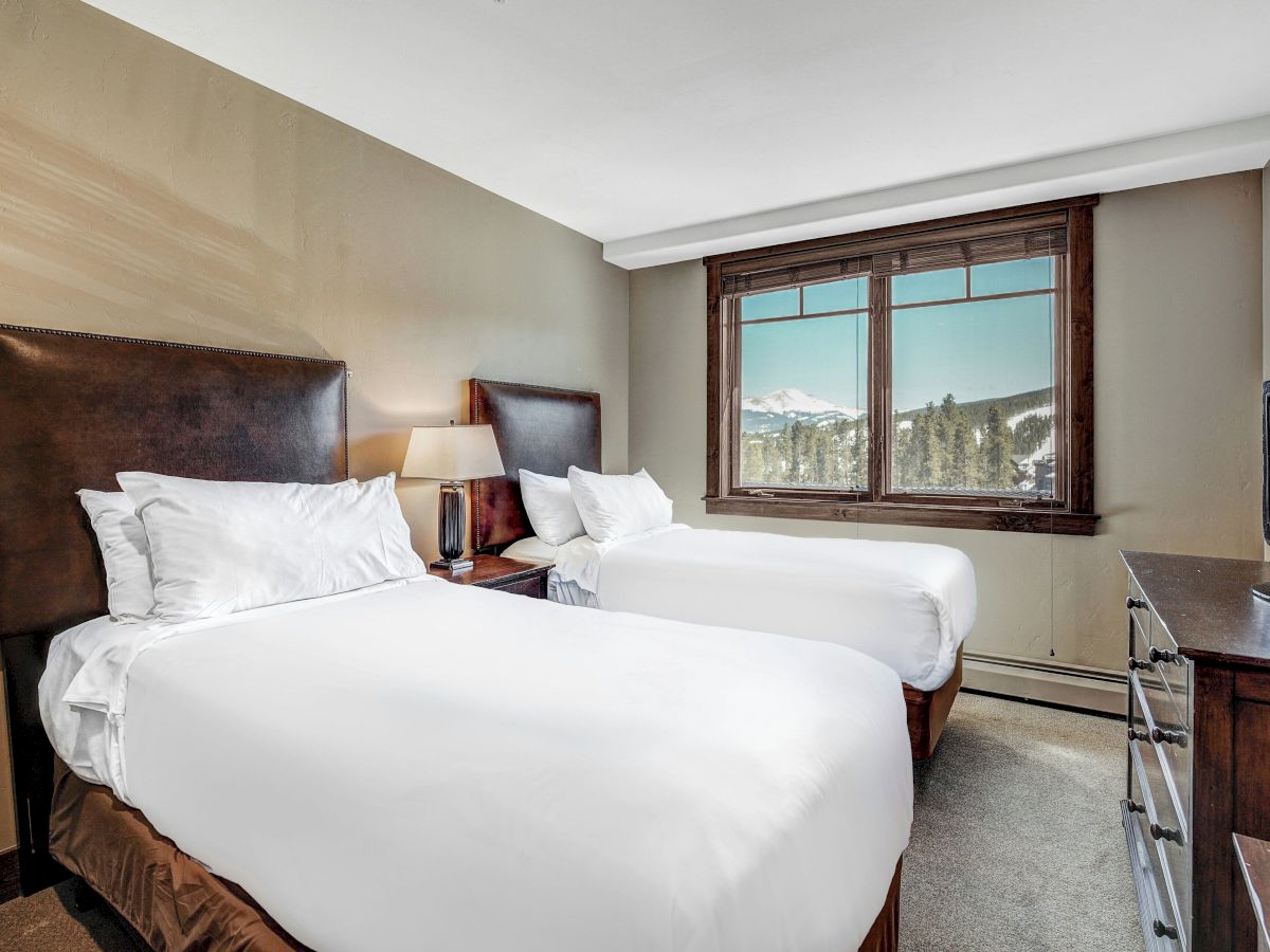 Cozy hotel bedroom with a large bed, bedside lamp, big window showing snowy mountains, and a TV on the dresser.