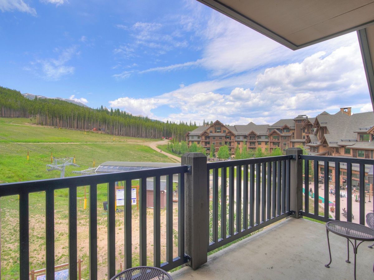 A sunny mountain resort view from a balcony, looking over a green ski slope, pine trees, and chalet buildings under a blue sky.