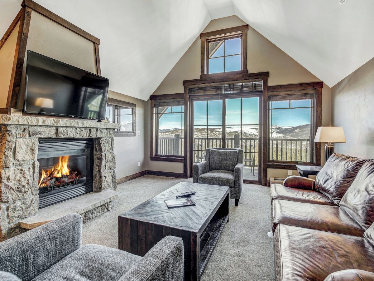 Cozy living room with stone fireplace, TV, sofa and armchairs, and large windows overlooking mountains, all under vaulted ceilings.