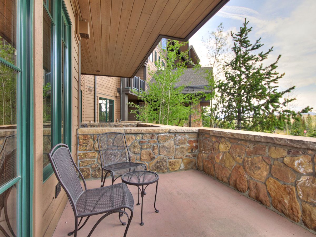 Outdoor balcony patio with two metal chairs, a stone wall, and green trees under an overhang.