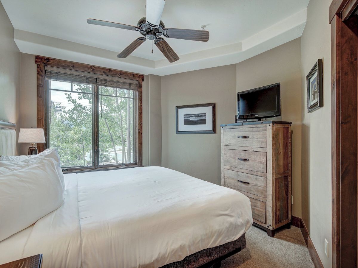 A bright bedroom with a ceiling fan, large window, bed, and a dresser holding a flat-screen TV.