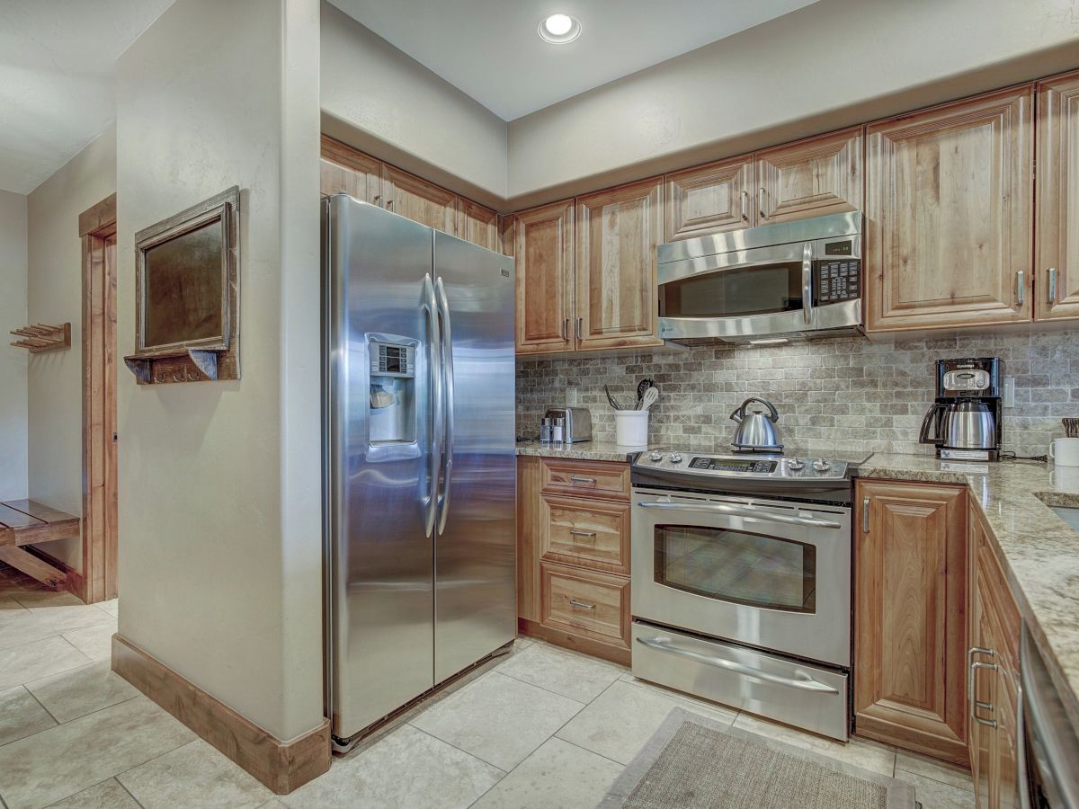 A bright kitchen with wood cabinets, stainless refrigerator, built-in microwave, gas range/oven, and a tiled backsplash with kettle and coffee makers.