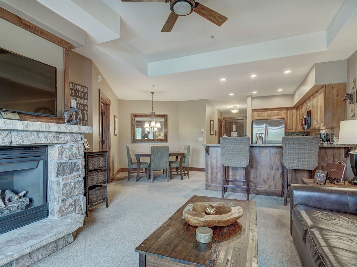 Bright living room with stone fireplace, TV, ceiling fan, dining table, and wood kitchen cabinets.