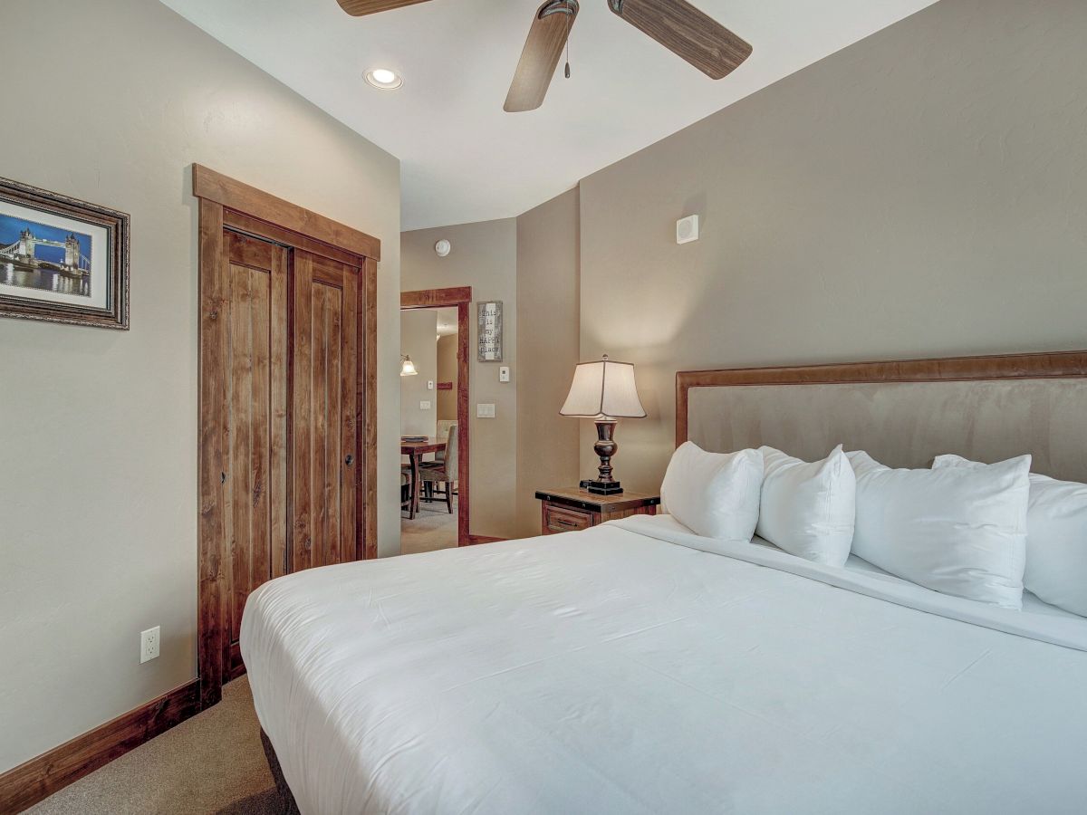 A bright bedroom with a large bed, white bedding, a wooden door, framed wall art, and a ceiling fan casting light on the wall.