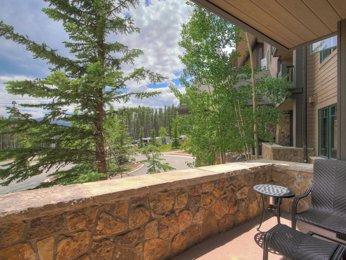 A patio with stone wall and chairs overlooks a quiet road, lush green trees, and a row of lodge-like buildings under cloudy skies.