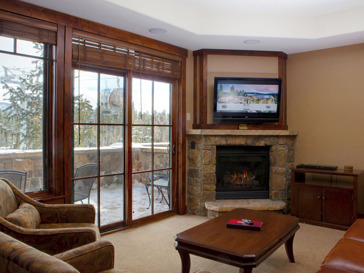 A cozy living room with a stone fireplace, TV, large windows showing snowy trees, wood blinds, and seating.
