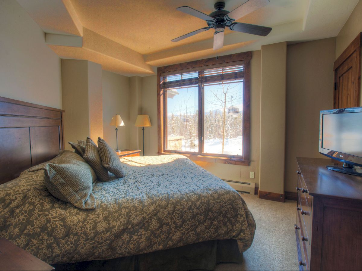 A cozy bedroom with a large bed, ceiling fan, window with snowy trees view, and a mounted TV on a dresser.
