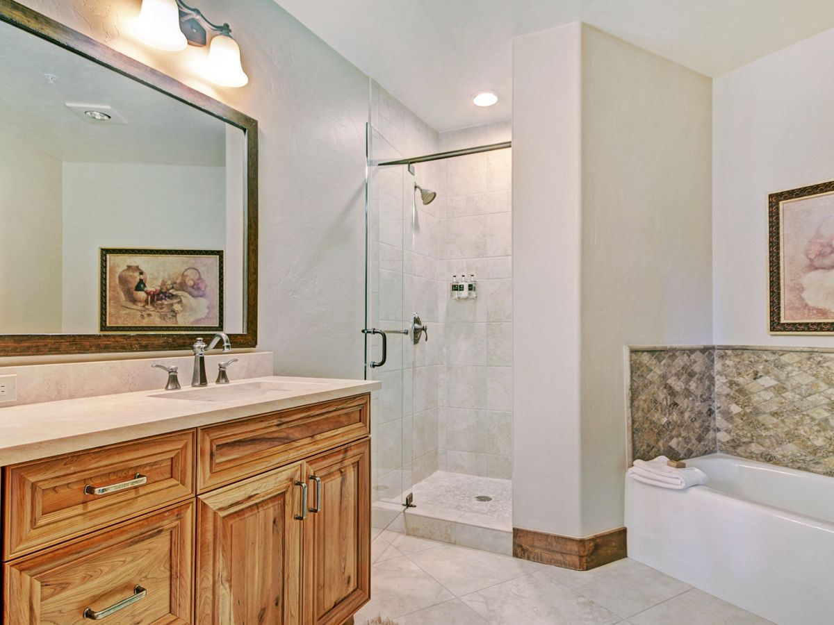 A bright bathroom with wooden vanity, glass shower, tiled floor, bathtub, and framed wall art.