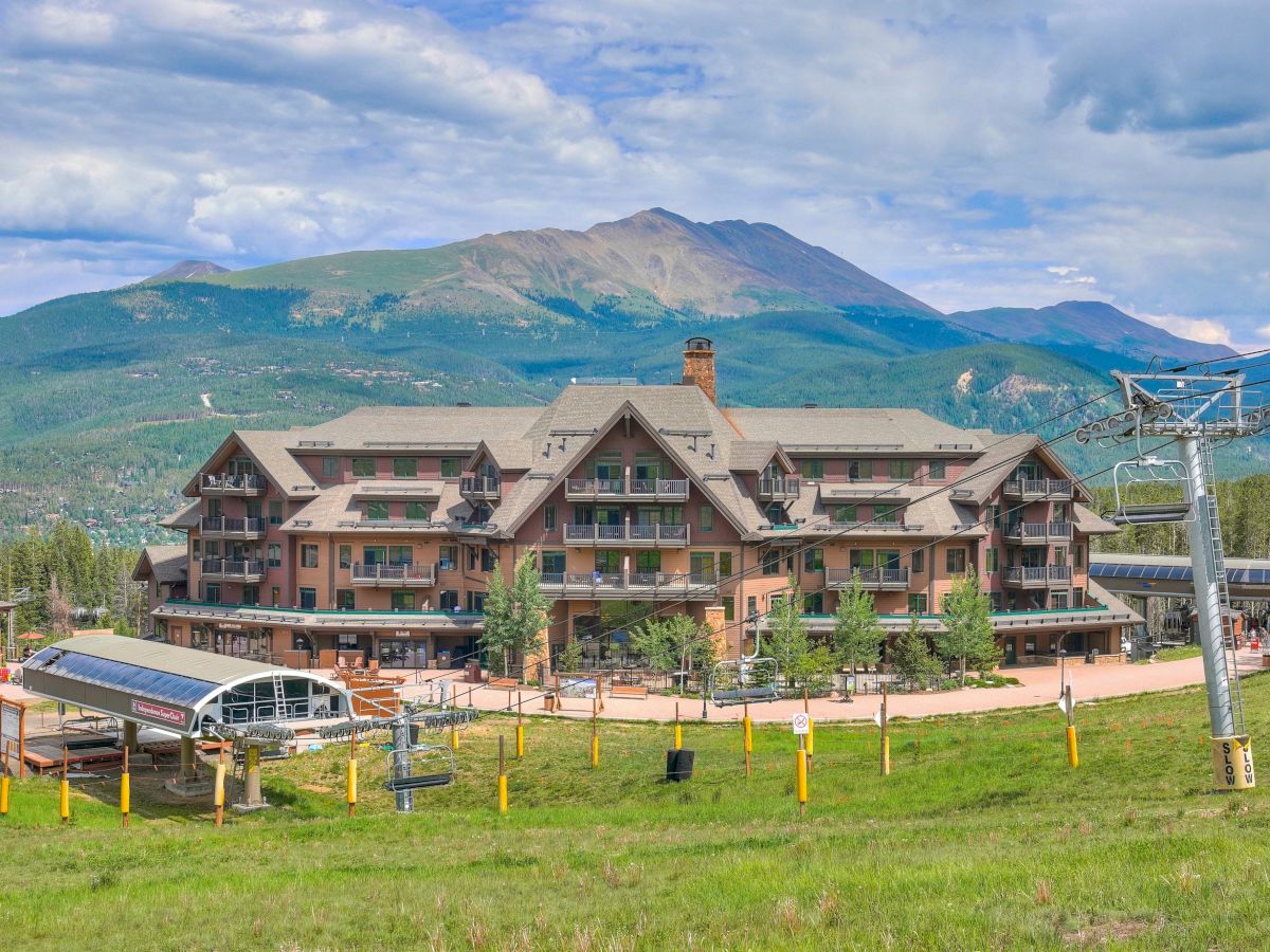 A mountain ski resort lodge with chairlift equipment sits beneath green peaks and a cloudy sky.