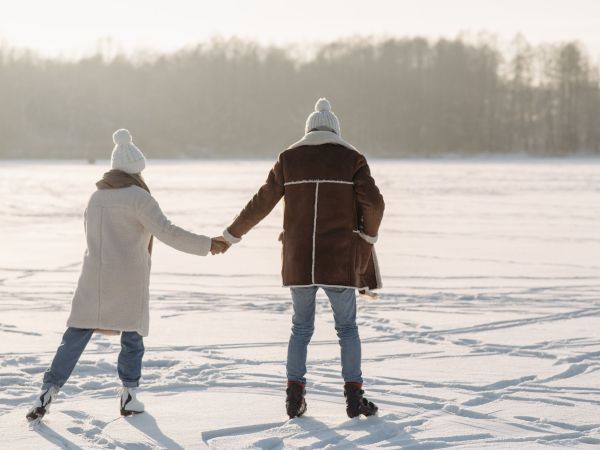 Two people in winter coats and knit hats hold hands while skating or walking on a snowy, frozen landscape.
