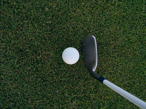 A white golf ball sits on green grass beside a golf club (driver) head, ready for a swing.