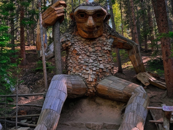 A large wooden animal statue with a slanted hat and paws, nestled in a forest of tall trees and dappled sunlight.