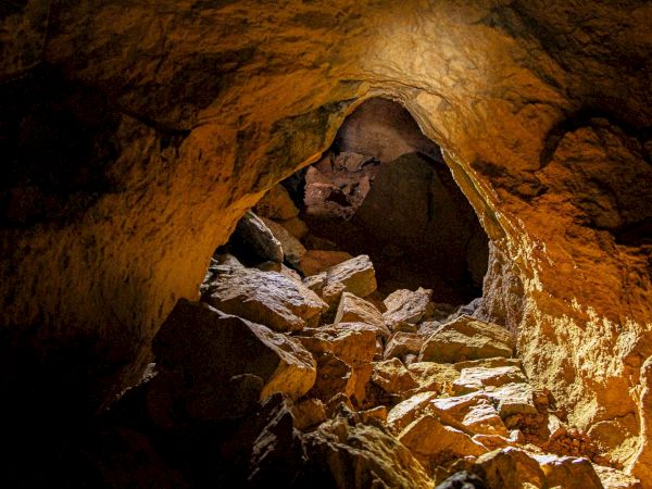 A rocky cave passage with crumbling golden walls, rubble on the floor, and warm light shining through the opening.