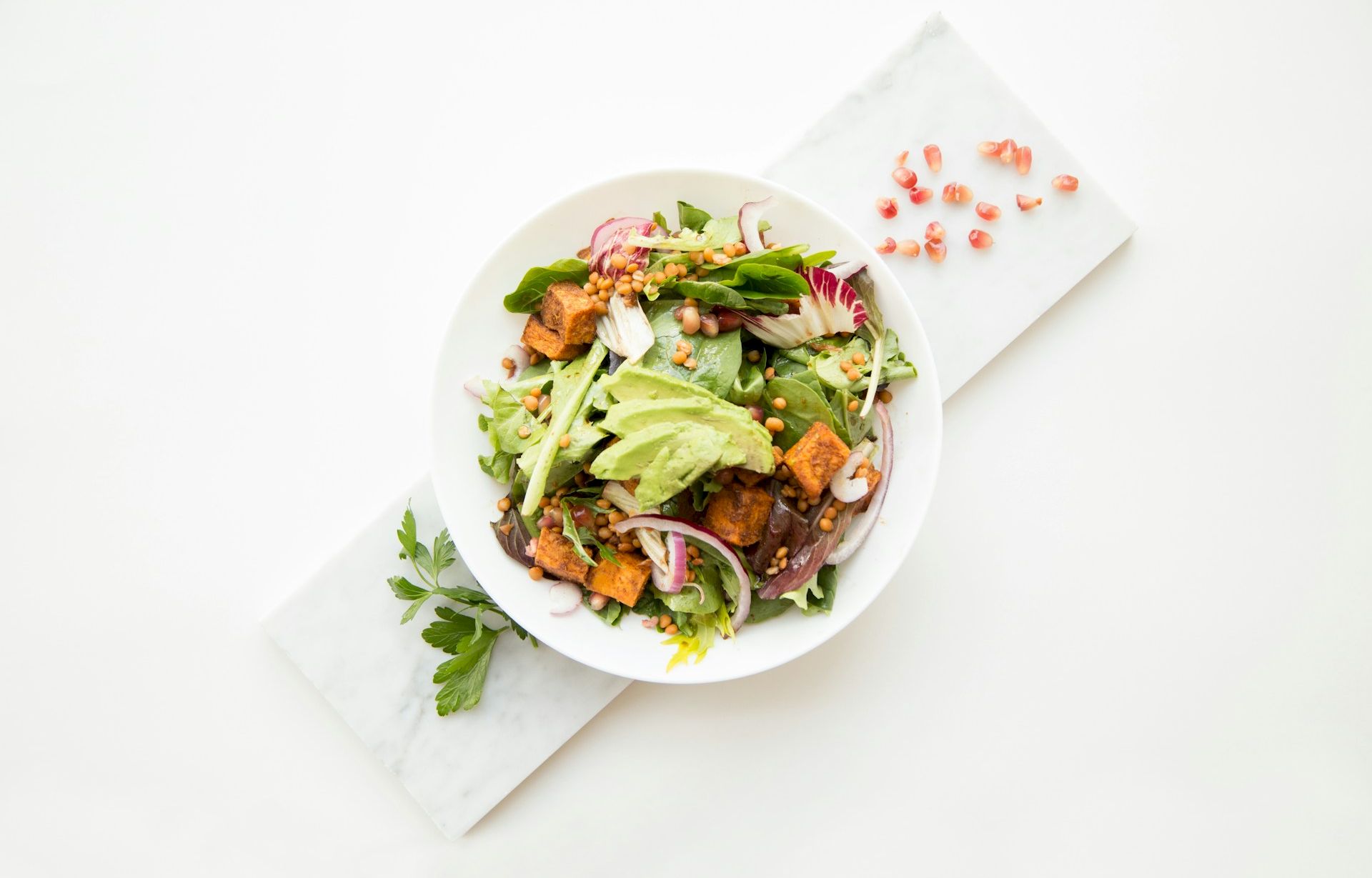 A bowl of fresh salad with leafy greens, lentils, avocado slices, crunchy croutons, and red onion on a white marble surface.