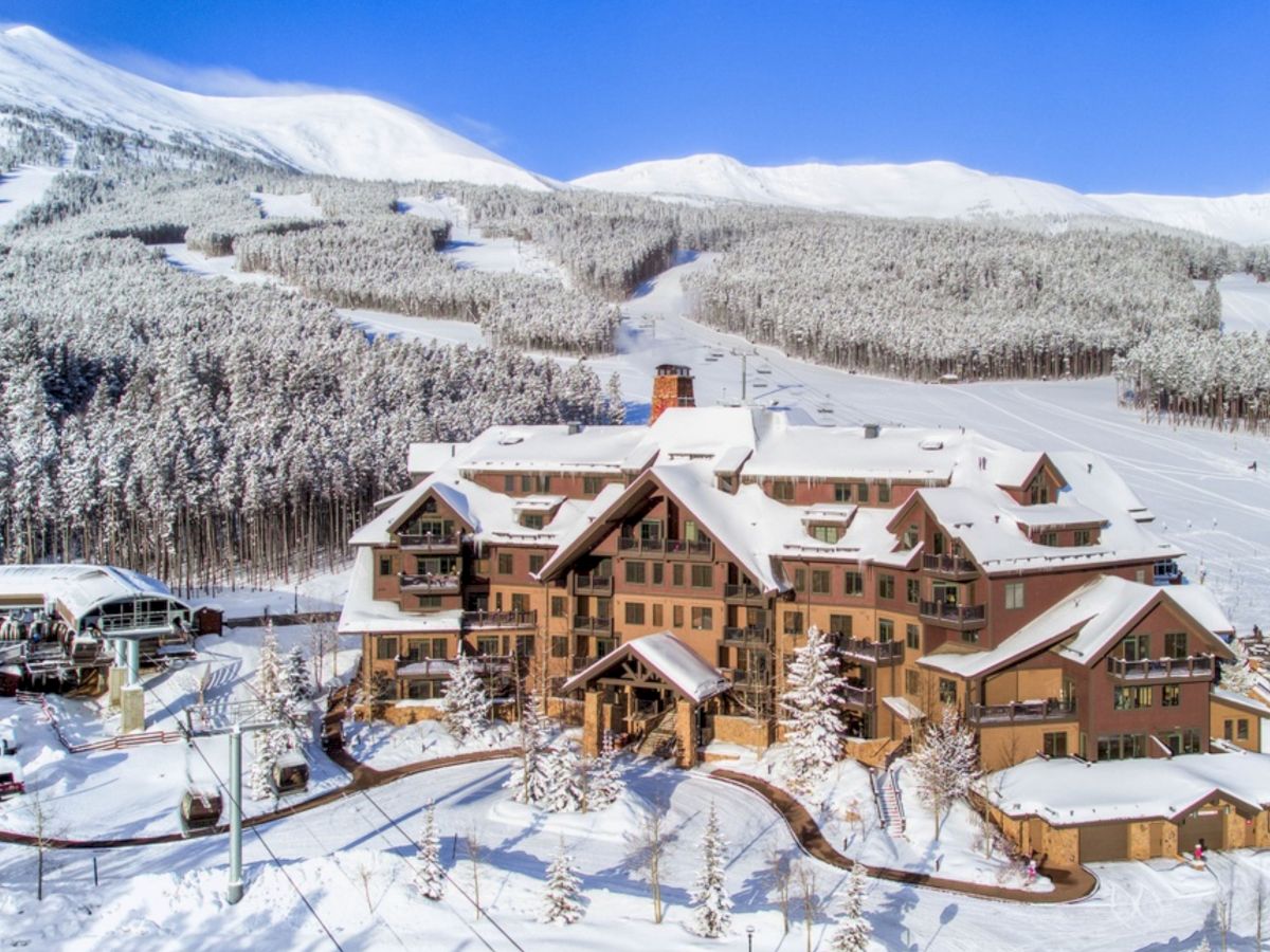 A snowy mountain ski resort with a lodge, winding road, and snowy forests under a bright blue sky.