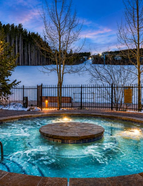 An outdoor hot tub at dusk with glowing lights, snow-covered ground, bare trees, and a forested yard behind a metal fence.