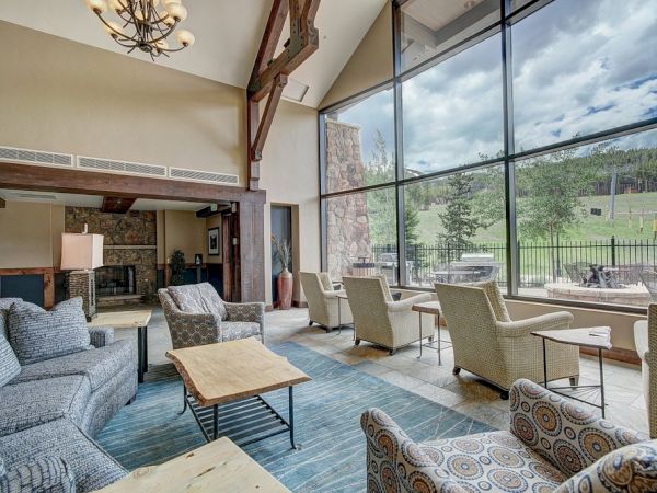 A bright lodge-style living room with sofas and patterned chairs, a fireplace, and large floor-to-ceiling windows by greenery.