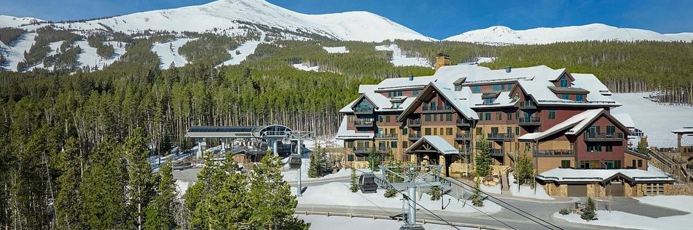 Snowy ski resort with a lodge and gondola lift, nestled in pine forest under bright mountains.