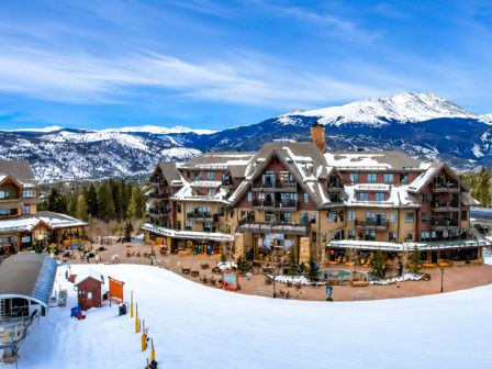 Snowy mountain resort with a large lodge/hotel, outdoor patio and activities, framed by snowy peaks under a blue sky.
