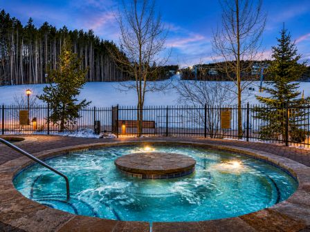 A night outdoor hot tub with bubbling water, stone deck lights, snowy grounds, bare trees, and a fence under a blue sky.