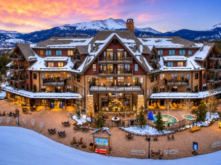 A snowy mountain resort courtyard with a large lodge, lit windows, and an outdoor pool under colorful dusk skies.