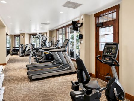 A bright fitness room with treadmills and exercise equipment, large windows, and a wall-mounted control panel near the door.