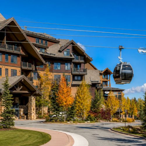 A ski resort lodge with balconies, autumn trees, and a cable-hauled gondola cabin against a blue sky.