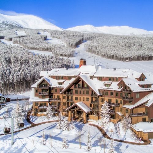 A snowy ski resort with a lodge, winding roads, and pine forests under bright blue mountains.