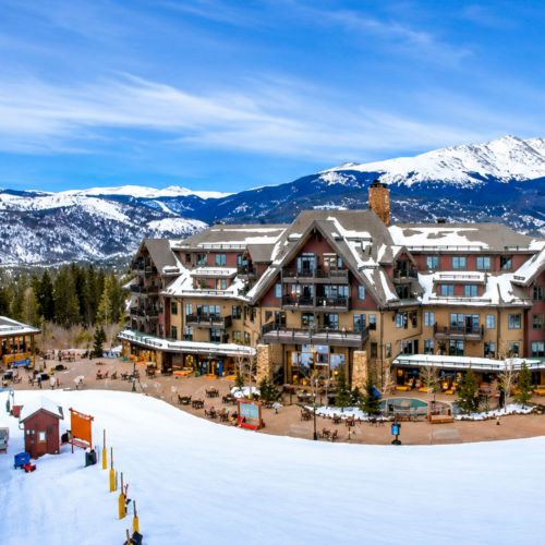 A snowy ski resort hotel sits in the foreground, with pine trees and a bright-blue sky above snowy mountain peaks.