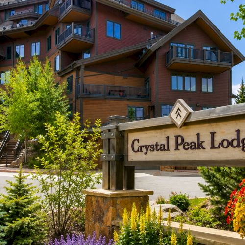 A sunny view of Crystal Peak Lodge with a landscaped entrance sign, trees, and a brick apartment-style building with balconies.