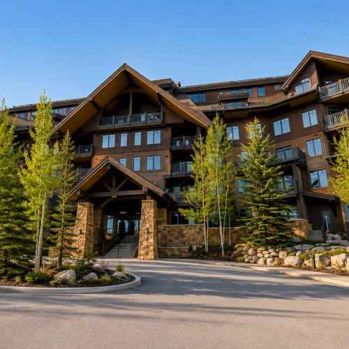 A modern multi-story apartment building with balconies, stone entrance, and fresh green trees lining a sunny driveway.