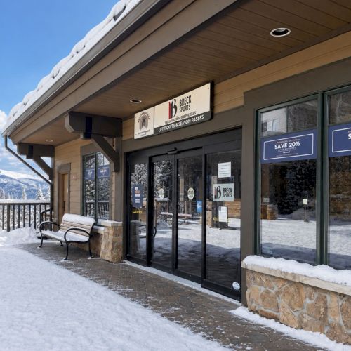 Snowy ski resort storefront with glass doors and signs reading &ldquo;Save 20%&rdquo; and &ldquo;Brecken Sports,&rdquo; outside in winter.