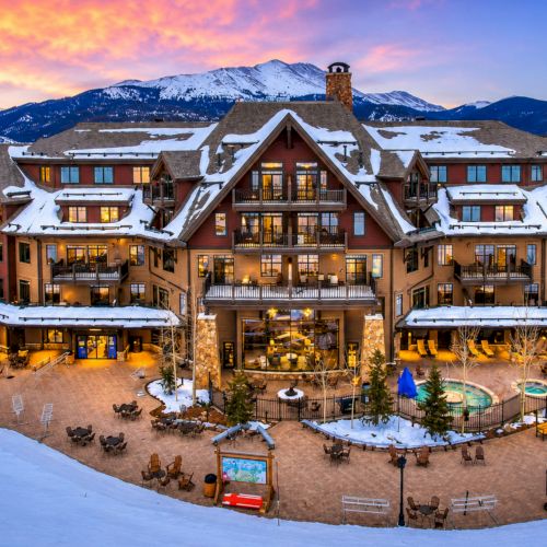 A snowy alpine resort lodge with balconies, warm lights, a courtyard pool, and mountains in the colorful sunset sky.