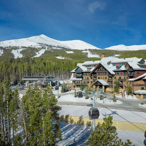 A snowy ski resort lodge sits by a chairlift with pine trees and distant mountains under a clear blue sky.