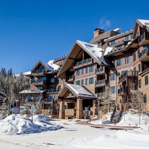 A snowy mountain lodge with multi-story balconies and peaked roofs under a bright blue winter sky, surrounded by trees and snow.