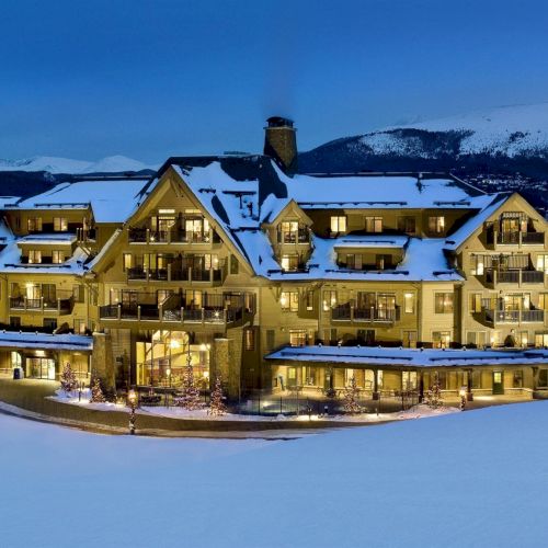 A snow-covered alpine hotel with lit windows and balconies sits in the mountains at dusk, surrounded by fresh white fields.