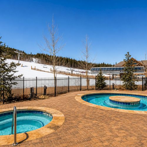 Outdoor resort pool with jets, patio tiles, distant ski slope and cabins, pine trees, and clear blue sky.