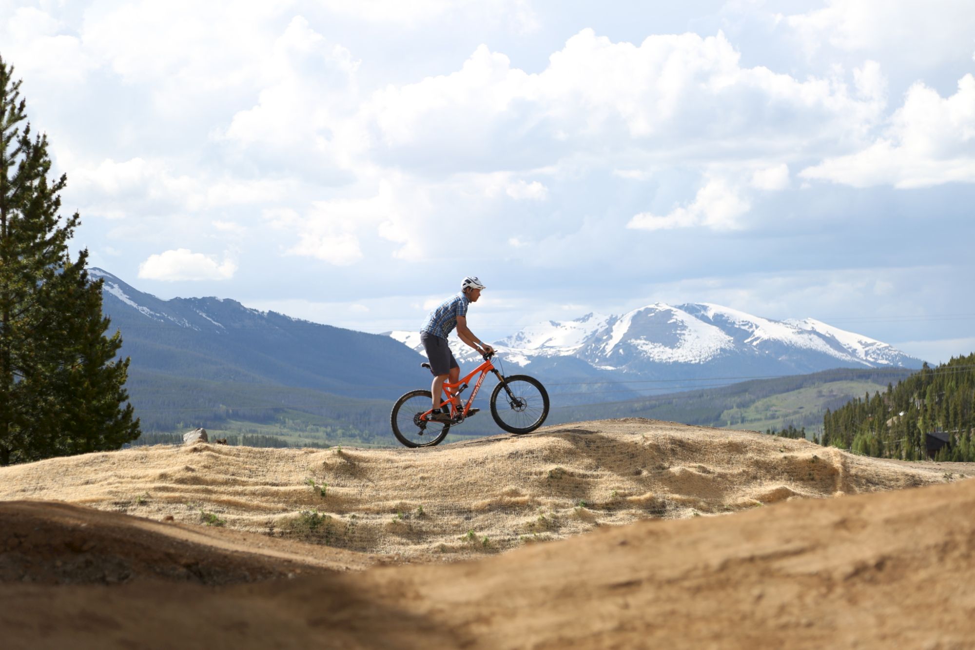 A mountain biker in a helmet rides along a dirt track with snowy peaks and pine forests in the background, chasing adventure.
