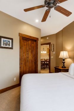 A cozy hotel bedroom with a king bed, ceiling fan, bedside lamp, wall art, TV on a dresser, and an open doorway.