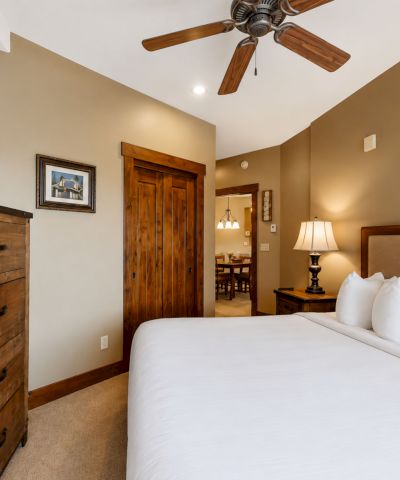 A cozy hotel bedroom with a king bed, ceiling fan, bedside lamp, wall art, TV on a dresser, and an open doorway.