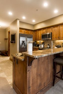 A cozy home kitchen with wooden cabinets, granite island, stainless appliances, chairs, and a large wall clock.