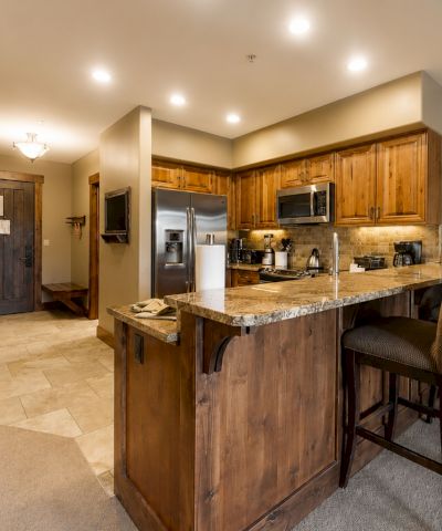 A cozy home kitchen with wooden cabinets, granite island, stainless appliances, chairs, and a large wall clock.